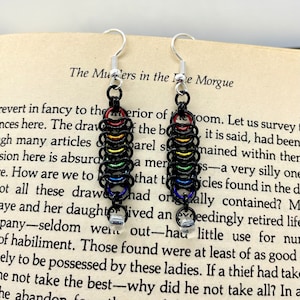 May include: A pair of rainbow chainmail earrings with silver hooks and clear beads. The earrings are hanging from a book with the text "The Murders in the Rue Morgue".