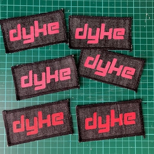 May include: Six black rectangular patches with the word "dyke" in red block letters. The patches are arranged on a green cutting mat. The patches are made of a dark fabric with a visible weave.