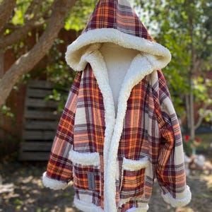 Op de afbeelding: Een getailleerd jack met capuchon en een witte sherpa voering. Het jack heeft een bruin, oranje en wit geruit patroon. Het jack heeft twee zakken aan de voorkant.