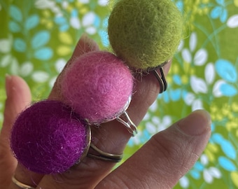 May include: Three felt ball rings in purple, pink, and green. The rings are on a finger.