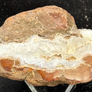 Op de afbeelding: Een natuurlijke, ongesneden agaatsteen met een gelaagde uitstraling. Het bovenste gedeelte is lichtbruin van kleur, overgaand in een witte en doorschijnende kern en een roodbruine basis. De steen wordt op een transparante standaard getoond.