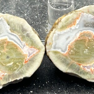 May include: Two halves of a geode with a white, green, and orange agate pattern. The geode is cut in half and polished to reveal the intricate patterns inside.