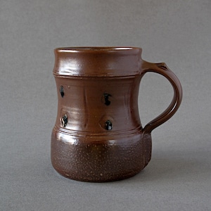 May include: A brown ceramic mug with a handle. The mug has a textured, speckled base and a smooth, glossy upper portion. Dark, irregular markings are spaced around the body of the mug. The handle is curved and integrated into the mug's design.