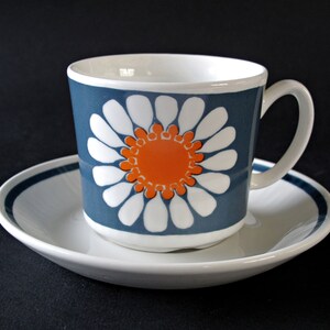 K&ouml;nnte beinhalten: Eine wei&szlig;e Teetasse und Untertasse aus Keramik mit einem blauen und orangefarbenen G&auml;nsebl&uuml;mchenmuster. Die Tasse hat einen Henkel und die Untertasse einen blauen Rand.