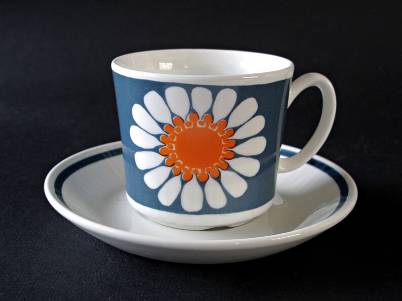 K&ouml;nnte beinhalten: Eine wei&szlig;e Teetasse und Untertasse aus Keramik mit einem blauen und orangefarbenen G&auml;nsebl&uuml;mchenmuster. Die Tasse hat einen Henkel und die Untertasse einen blauen Rand.