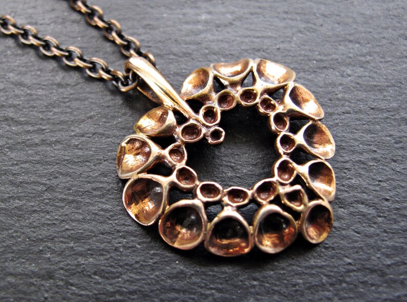 May include: A bronze pendant necklace with a circular design featuring a repeating pattern of small, rounded shapes. The pendant is attached to a black chain.