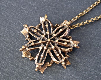 May include: A gold-toned pendant necklace featuring a detailed snowflake design with a textured finish. The snowflake has a circular center with radiating lines and small dots.