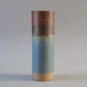 May include: A tall, cylindrical ceramic vase with a textured surface. The top section is a deep reddish-brown, transitioning to a darker brown band, then a blue-green middle, and a tan base. The vase is approximately 25 cm tall.