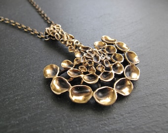 May include: A gold-toned pendant necklace featuring a large, abstract, organic-shaped design. The pendant is crafted from numerous small, rounded, overlapping pieces, creating a textured, three-dimensional effect.