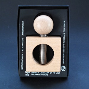May include: A wooden nutcracker with a screw mechanism. The nutcracker is in a black box with the text "NØDDEKNÆKKER - NUTCRACKERS - NUSSKNACKER" and "MADE IN DENMARK BY PP LINE DK 8900 RANDERS" and "DESIGN: RUD THYGESEN & JOHNNY SØRENSEN".