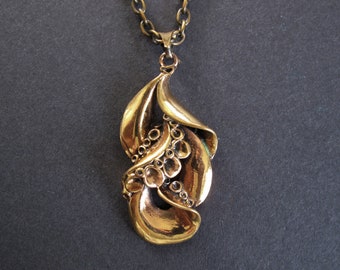 May include: A gold-toned pendant necklace with an abstract, organic design. The pendant features flowing, leaf-like shapes and a cluster of small, circular details. The chain is a matching gold color, creating a cohesive and elegant piece of jewelry.