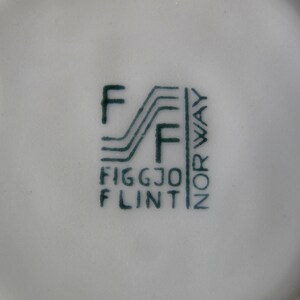 K&ouml;nnte beinhalten: Nahaufnahme einer gr&uuml;nen und wei&szlig;en Keramikmarke mit dem Text "Figgjo Flint" und "Norwegen" in einer stilisierten Schrift.