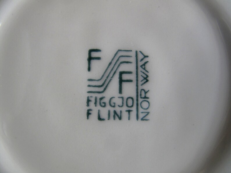 K&ouml;nnte beinhalten: Nahaufnahme einer gr&uuml;nen und wei&szlig;en Keramikmarke mit dem Text "Figgjo Flint" und "Norwegen" in einer stilisierten Schrift.