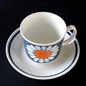 K&ouml;nnte beinhalten: Eine wei&szlig;e Teetasse und Untertasse mit einem blauen und orangefarbenen G&auml;nsebl&uuml;mchen-Design. Die Tasse hat ein wei&szlig;es Inneres und einen blauen Rand. Die Untertasse hat einen blauen Rand und eine wei&szlig;e Mitte.