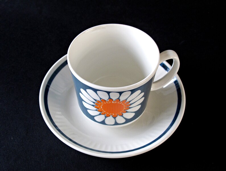 K&ouml;nnte beinhalten: Eine wei&szlig;e Teetasse und Untertasse mit einem blauen und orangefarbenen G&auml;nsebl&uuml;mchen-Design. Die Tasse hat ein wei&szlig;es Inneres und einen blauen Rand. Die Untertasse hat einen blauen Rand und eine wei&szlig;e Mitte.