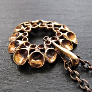 May include: A bronze pendant with a circular design featuring a repeating pattern of small, rounded shapes. The pendant is attached to a bronze chain.