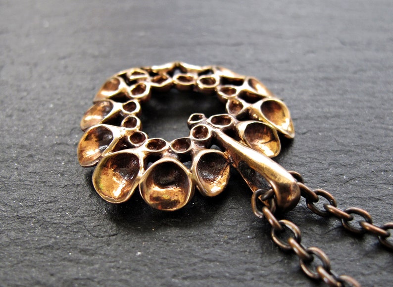 May include: A bronze pendant with a circular design featuring a repeating pattern of small, rounded shapes. The pendant is attached to a bronze chain.