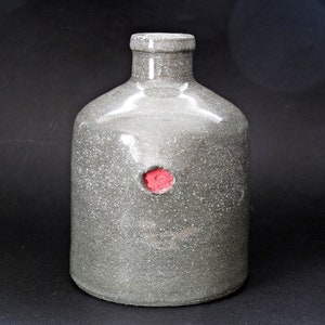 以下が含まれることがあります： 斑点のある質感と赤いシールが付いた灰色のセラミックボトル。ボトルは丸みを帯びたボディと細い首を持っています。セラミックボトルは装飾品です。
