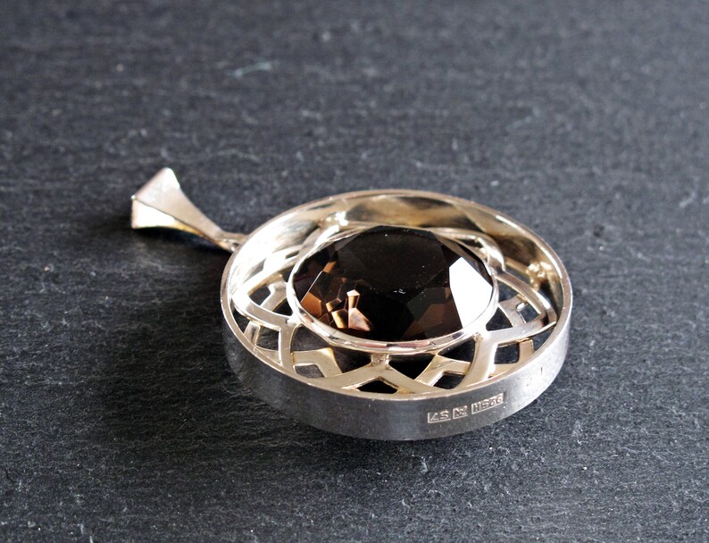 May include: A silver pendant featuring a dark brown gemstone set within a geometric design. The pendant has a bail and is hallmarked "925" on the reverse.