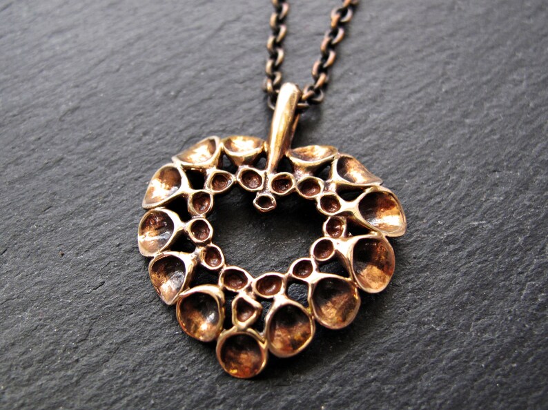 May include: A bronze pendant in the shape of a heart, with a textured design of small, rounded shapes. The pendant is attached to a chain.