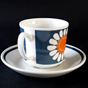 K&ouml;nnte beinhalten: Eine wei&szlig;e Teetasse und Untertasse aus Keramik mit einem blauen und orangefarbenen G&auml;nsebl&uuml;mchenmuster. Die Tasse hat einen abgerundeten Griff und die Untertasse hat einen blauen Streifen am Rand.