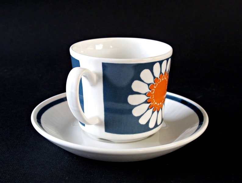 K&ouml;nnte beinhalten: Eine wei&szlig;e Teetasse und Untertasse aus Keramik mit einem blauen und orangefarbenen G&auml;nsebl&uuml;mchenmuster. Die Tasse hat einen abgerundeten Griff und die Untertasse hat einen blauen Streifen am Rand.