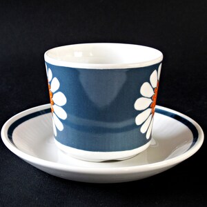 K&ouml;nnte beinhalten: Eine blaue und wei&szlig;e Kaffeetasse aus Keramik mit einem G&auml;nsebl&uuml;mchenmuster und orangefarbenen Akzenten. Die Tasse steht auf einer wei&szlig;en Untertasse mit einem blauen Band.