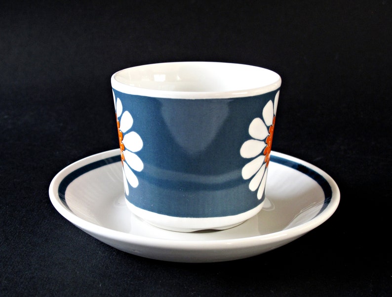K&ouml;nnte beinhalten: Eine blaue und wei&szlig;e Kaffeetasse aus Keramik mit einem G&auml;nsebl&uuml;mchenmuster und orangefarbenen Akzenten. Die Tasse steht auf einer wei&szlig;en Untertasse mit einem blauen Band.
