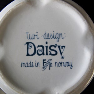 K&ouml;nnte beinhalten: Wei&szlig;e Keramiktasse mit blau-wei&szlig;em Design. Die Tasse tr&auml;gt die Aufschrift "turi-design: Daisy made in F/F norway".