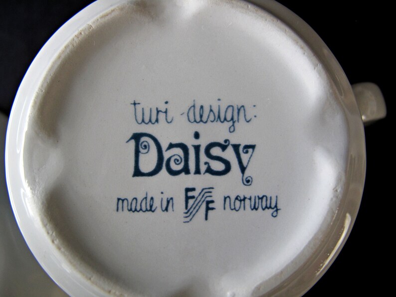 K&ouml;nnte beinhalten: Wei&szlig;e Keramiktasse mit blau-wei&szlig;em Design. Die Tasse tr&auml;gt die Aufschrift "turi-design: Daisy made in F/F norway".