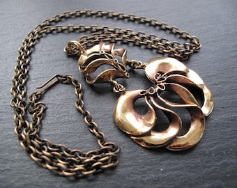 May include: A vintage-style bronze necklace with a detailed pendant. The pendant features layered, curved shapes and is suspended from a matching chain. The necklace has a hook clasp and is displayed on a dark surface.