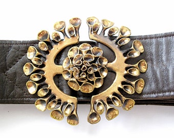 May include: A gold-toned metal belt buckle with a unique, organic design. The buckle features a central circular motif surrounded by intricate, branching patterns.