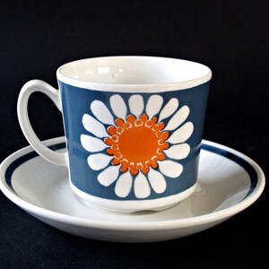K&ouml;nnte beinhalten: Eine wei&szlig;e Teetasse und Untertasse aus Keramik mit einem blauen und orangefarbenen Blumendesign. Die Tasse hat eine wei&szlig;e Margerite mit einer orangefarbenen Mitte auf blauem Hintergrund. Die Untertasse hat einen blauen Rand.