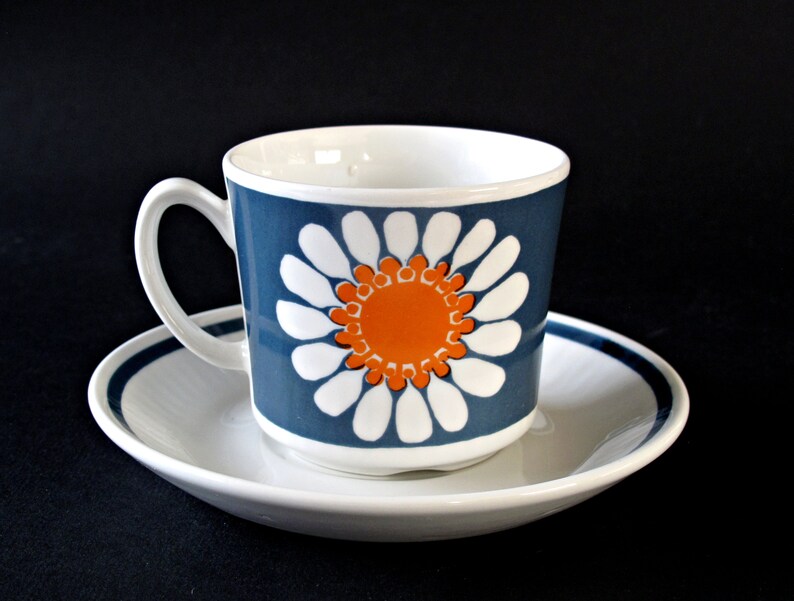 K&ouml;nnte beinhalten: Eine wei&szlig;e Teetasse und Untertasse aus Keramik mit einem blauen und orangefarbenen Blumendesign. Die Tasse hat eine wei&szlig;e Margerite mit einer orangefarbenen Mitte auf blauem Hintergrund. Die Untertasse hat einen blauen Rand.