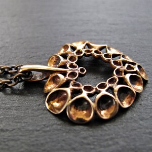 May include: A bronze pendant necklace with a circular design featuring a repeating pattern of small, rounded shapes. The pendant is attached to a chain.
