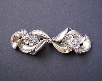May include: A silver brooch with a unique, abstract design featuring swirling, organic shapes and textured details.