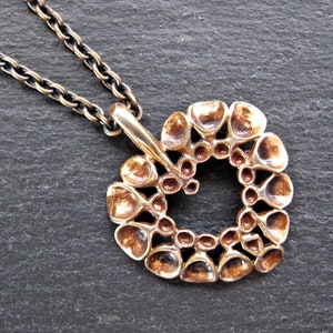 May include: A bronze pendant necklace with a circular design featuring a series of interconnected, rounded shapes. The pendant has a textured, organic look.