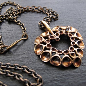 May include: A bronze necklace with a large, intricate pendant shaped like a heart. The pendant is made up of many small, rounded shapes that create a textured, organic look.