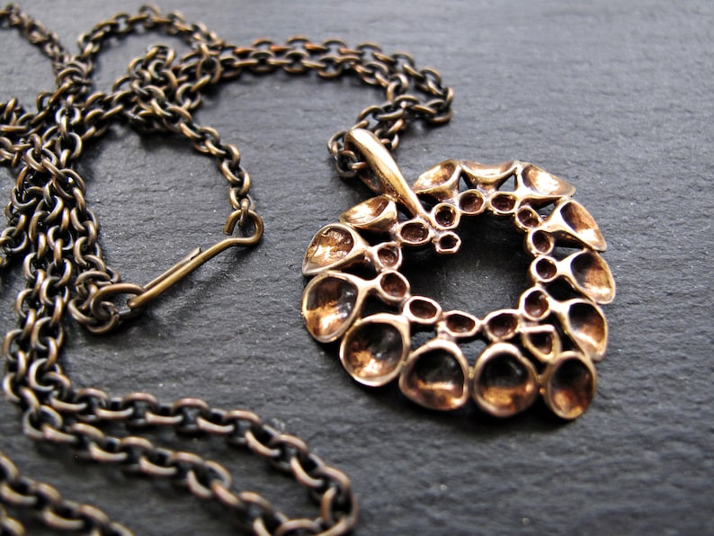 May include: A bronze necklace with a large, intricate pendant shaped like a heart. The pendant is made up of many small, rounded shapes that create a textured, organic look.