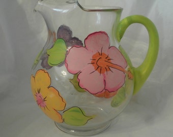 May include: A clear glass pitcher with a green handle, decorated with hand-painted pink, yellow, and green hibiscus flowers.