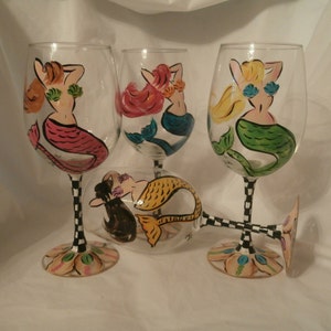 May include: Four hand-painted wine glasses featuring mermaid designs. Each glass has a different mermaid with colorful hair and tails. The stems have a black and white checkered pattern, and the bases are decorated with floral designs.