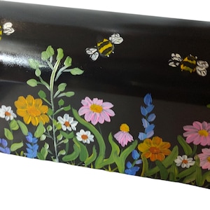May include: Black mailbox with a hand-painted floral design featuring yellow, pink, and white flowers, green leaves, and blue accents. Several painted bees are scattered across the top, creating a whimsical garden theme.