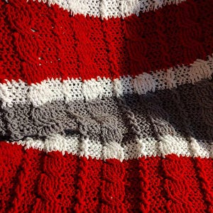 Puede incluir: Primer plano de una manta de ganchillo hecha a mano con un patrón texturizado. La manta presenta franjas alternas de color rojo, blanco y gris. Las franjas rojas tienen un diseño de punto de cable, mientras que las franjas blancas y grises tienen una puntada más simple.