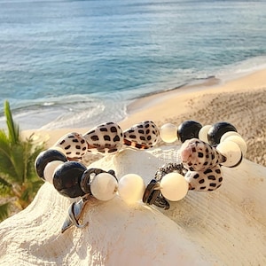 May include: A bracelet made with black and white beads, seashells, and a silver charm. The bracelet is on a white seashell against a background of a beach and ocean.