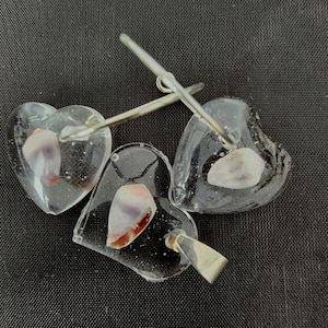 May include: Three clear glass heart-shaped pendants with silver jump rings. Each pendant contains a different seashell with brown, white, and pink colors.