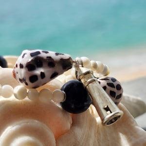 May include: A close-up of a silver whistle charm on a necklace with white and black shell beads. The necklace is resting on a white seashell.