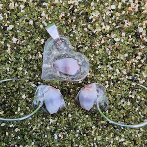 May include: A set of three heart-shaped resin jewelry pieces with seashells embedded inside. The necklace features a larger heart with a pink and white seashell, while the earrings feature smaller hearts with white seashells.