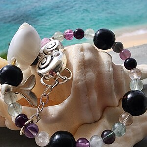 May include: A bracelet made with black and green beads, purple and pink beads, and a silver charm. The bracelet is on a seashell.