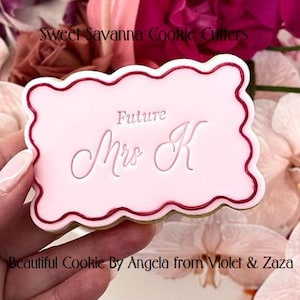 May include: Pink cookie decorated with a red border and the text "Future Mrs. K" in white icing.