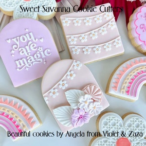 May include: An assortment of decorated sugar cookies with various designs, including a pink cookie with the text "you are magic", a pink cookie with a floral design, a pink cookie with a garland of flowers, and a rainbow cookie.